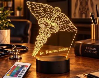 May include: An illuminated LED desk lamp featuring a glowing yellow caduceus symbol with wings and snakes. The acrylic panel is engraved with the text Dr. Francesca Richson. The lamp includes a black circular base and a remote control.