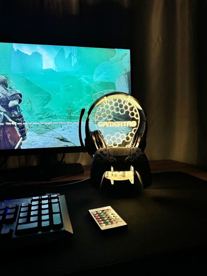 May include: A black gaming headset and controller stand with a clear acrylic base and a honeycomb design. The stand has a yellow light that illuminates the word "GAMERTAG" on the headset. The stand is on a black desk with a keyboard and a remote control.
