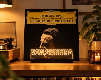 May include: A black and gold vinyl record display stand featuring the text NOW SPINNING in illuminated letters. The stand is positioned in front of a Frederic Chopin record cover by Arturo Benedetti Michelangeli, showcasing a portrait.