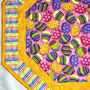 Può includere: Runner da tavolo pasquale trapuntato con sfondo rosa e disegni di uova colorate. Le uova presentano vari motivi e colori, tra cui strisce, pois e motivi floreali. Il runner ha un bordo giallo con accenti a righe e fantasia.