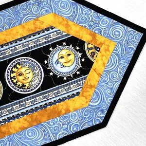 May include: A celestial-themed table runner with a black border. The design features a black center with smiling sun and moon faces, surrounded by stars. The runner has a gold and blue patterned border.