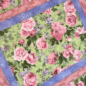 Può includere: Una trapunta floreale con un motivo di rose rosa e foglie verdi su sfondo verde chiaro. Ha un bordo lavanda e un bordo rosa a fantasia, creando un effetto a strati. La trapunta è probabilmente fatta a mano.