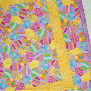 Può includere: Primo piano di una colorata trapunta a tema pasquale. La trapunta presenta un motivo di uova di Pasqua decorate in vari colori, tra cui rosa, giallo, arancione e blu, su uno sfondo lilla chiaro. I bordi sono bordati con tessuto giallo.