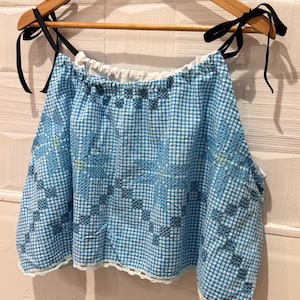 May include: A blue and white gingham crop top with black tie straps. The top has embroidered floral patterns in blue and black. The hemline is finished with a delicate white lace trim. The top is hanging on a wooden hanger.