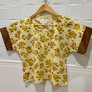 May include: A yellow floral print top with short sleeves and a square neckline. The top features a repeating pattern of yellow and orange flowers on a light yellow background. The short sleeves have a brown textured trim.