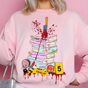 May include: Pink sweatshirt with a graphic design of a stack of books with a knife sticking through the top book. The books have titles related to true crime, such as "Murder Mystery", "Dead", "The Perfect Murder", and "Mysterious Death". There is a yellow police tape with the text "Police Line" and a red and white police siren. The number "5" is on a yellow sign.