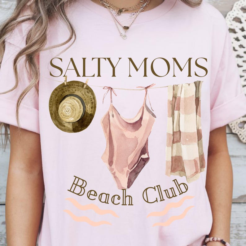 May include: A pink t-shirt with the text "Salty Moms Beach Club" in brown lettering. The text is surrounded by a watercolour illustration of a swimsuit, a towel, and a hat hanging on a clothesline.