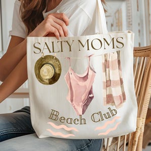 May include: A white canvas tote bag with the text "Salty Moms Beach Club" in brown lettering. The bag features a pink swimsuit, a brown hat, and a striped towel hanging on a clothesline.