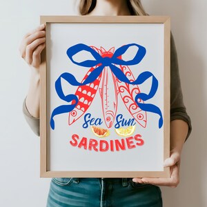 Puede incluir: Una impresi&oacute;n enmarcada con una cinta azul atada alrededor de tres sardinas rojas y blancas. El texto "Sea Sun Sardines" est&aacute; debajo de las sardinas, con rodajas de pomelo rojo y lim&oacute;n amarillo.