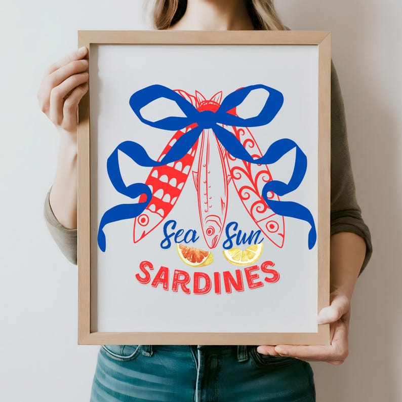 Puede incluir: Una impresi&oacute;n enmarcada con una cinta azul atada alrededor de tres sardinas rojas y blancas. El texto "Sea Sun Sardines" est&aacute; debajo de las sardinas, con rodajas de pomelo rojo y lim&oacute;n amarillo.
