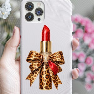 May include: A white smartphone case featuring a central graphic of a bright red lipstick tube. The base of the lipstick is wrapped in a large, detailed leopard print bow with a small, rectangular, clear crystal embellishment at the center.