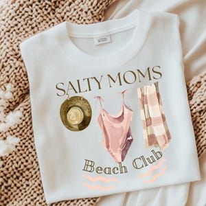 May include: White t-shirt with the text "Salty Moms Beach Club" in brown lettering. The design includes a brown hat, a pink swimsuit, and a striped towel.