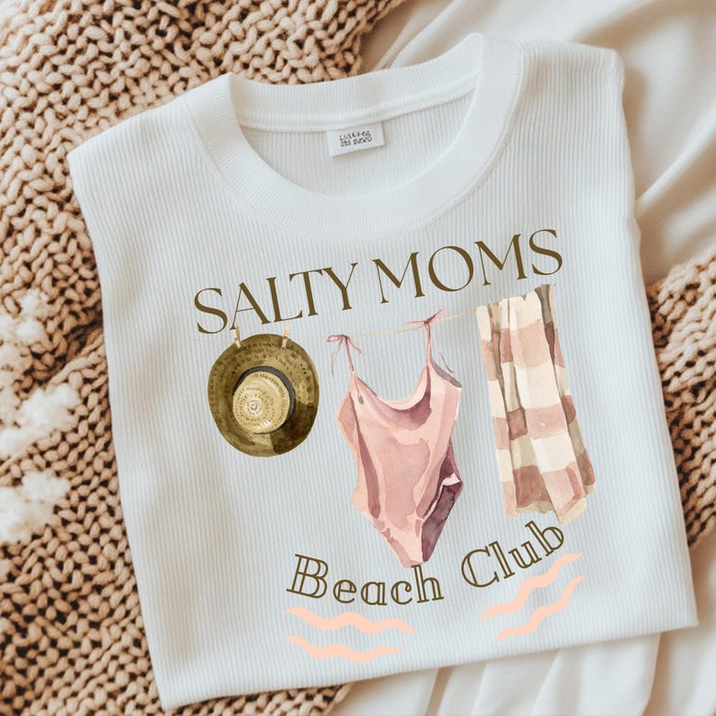 May include: White t-shirt with the text "Salty Moms Beach Club" in brown lettering. The design includes a brown hat, a pink swimsuit, and a striped towel.