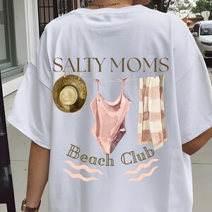 May include: White t-shirt featuring a graphic of a swimsuit, towel, and hat hanging on a clothesline. The text "Salty Moms Beach Club" is printed below the graphic.