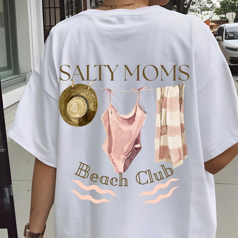 May include: White t-shirt featuring a graphic of a swimsuit, towel, and hat hanging on a clothesline. The text "Salty Moms Beach Club" is printed below the graphic.
