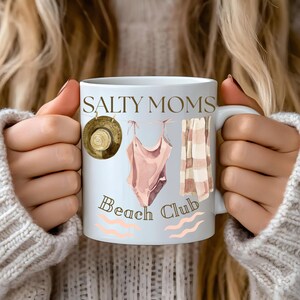 May include: White ceramic mug with a brown and white illustration of a swimsuit, towel, and hat. The text "Salty Moms Beach Club" is printed on the mug.