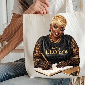 May include: Cream-colored tote bag featuring a graphic of a woman with blonde hair, wearing a black top with the text "In My CEO Era." The woman is writing in a notebook with a gold pen, with a gold laptop nearby.