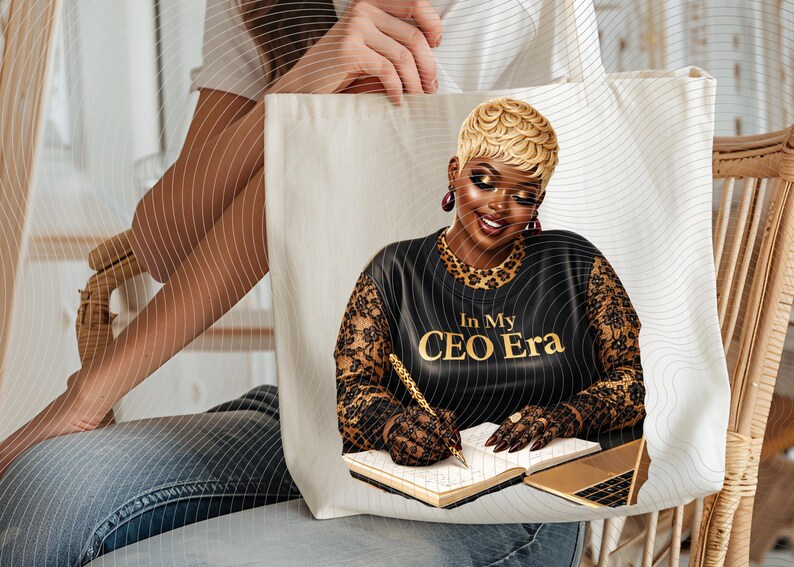 May include: Cream-colored tote bag featuring a graphic of a woman with blonde hair, wearing a black top with the text "In My CEO Era." The woman is writing in a notebook with a gold pen, with a gold laptop nearby.