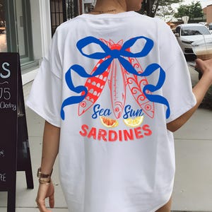 Puede incluir: Camiseta blanca con un lazo azul y tres peces rojos y blancos con el texto "Sea Sun Sardines" y una toronja y un lim&oacute;n.