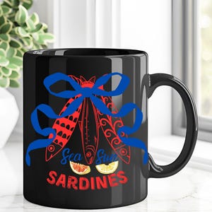 Puede incluir: Taza de caf&eacute; de cer&aacute;mica negra con una cinta azul y tres peces rojos y blancos con el texto "Sea Sun Sardines" en rojo y blanco.