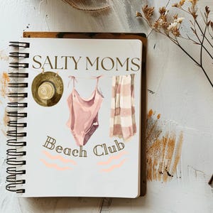 May include: A white notebook with a gold spiral binding. The notebook features a pink and white swimsuit, a straw hat, and a pink and white striped towel hanging from a clothesline. The text "Salty Moms Beach Club" is written in gold letters on the notebook.