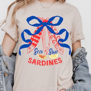 Puede incluir: Una camiseta beige claro con un lazo azul y tres peces rojos con dise&ntilde;os blancos y azules. El texto "Sea Sun Sardines" est&aacute; impreso en rojo y blanco debajo de los peces.