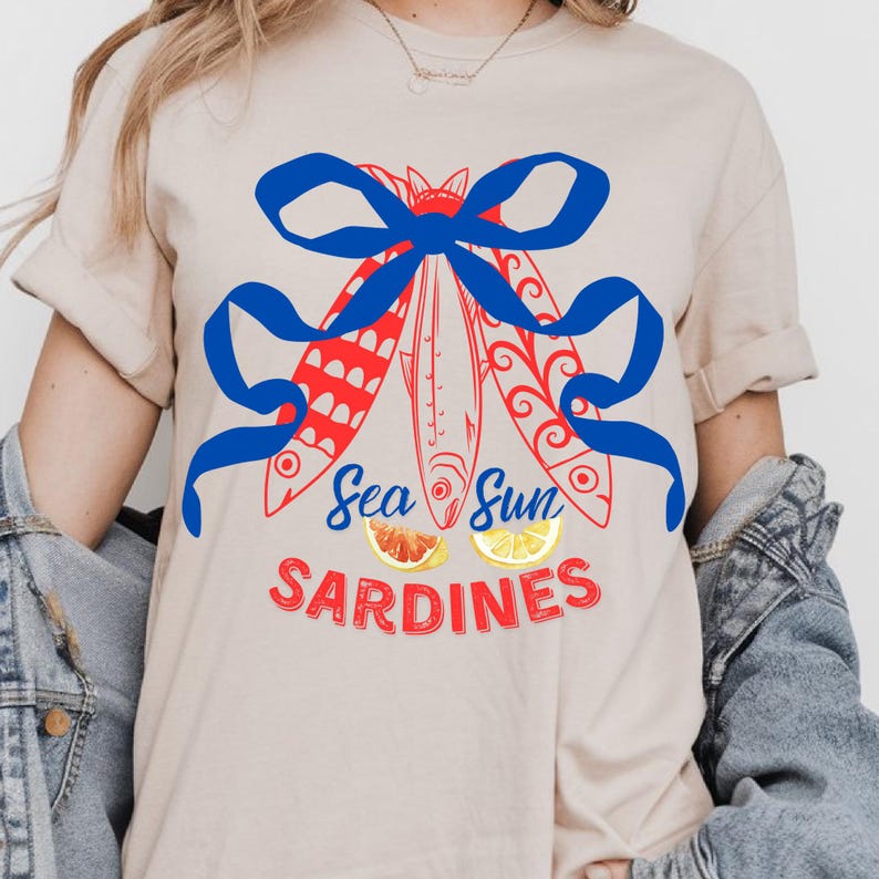 Puede incluir: Una camiseta beige claro con un lazo azul y tres peces rojos con dise&ntilde;os blancos y azules. El texto "Sea Sun Sardines" est&aacute; impreso en rojo y blanco debajo de los peces.