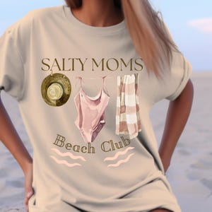 May include: A beige t-shirt with the text "Salty Moms Beach Club" printed on it. The text is in a brown font. There is a pink swimsuit, a striped towel, and a brown hat hanging on a line above the text.