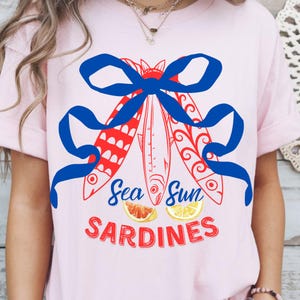 Puede incluir: Una camiseta rosa con un lazo azul y tres peces rojos y blancos con el texto "Sea Sun Sardines" y dos c&iacute;tricos.