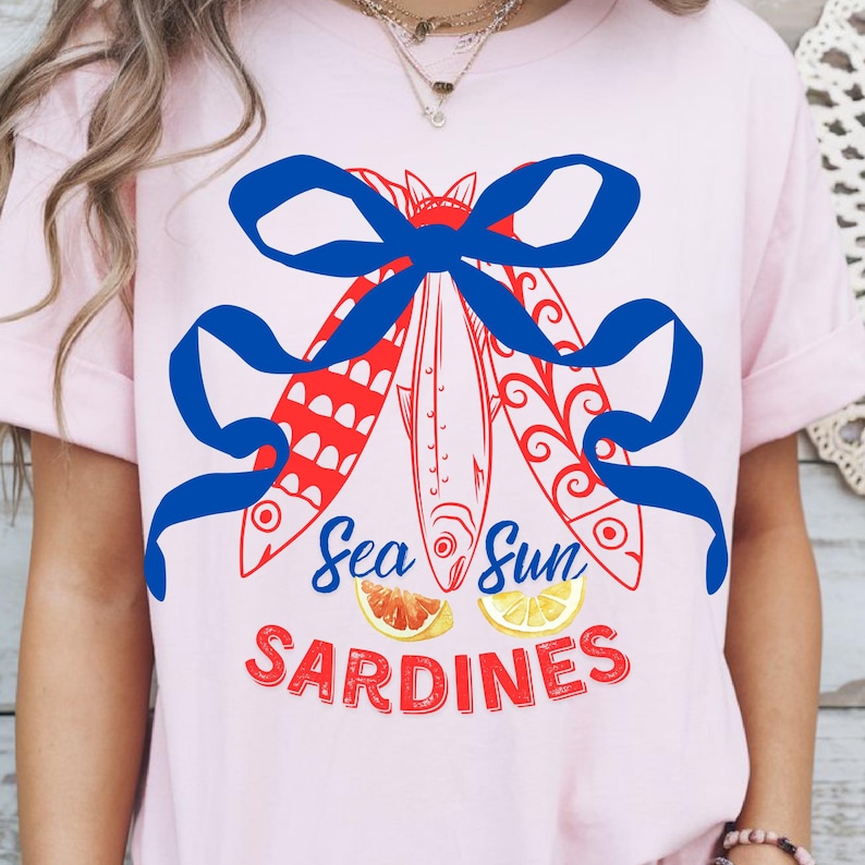 Puede incluir: Una camiseta rosa con un lazo azul y tres peces rojos y blancos con el texto "Sea Sun Sardines" y dos c&iacute;tricos.
