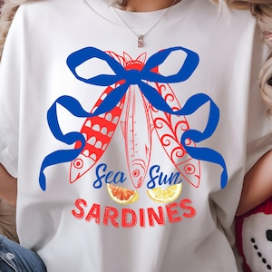 Puede incluir: Camiseta blanca con un lazo azul y una ilustraci&oacute;n de peces rojos y blancos. El texto "Sea Sun Sardines" est&aacute; impreso debajo de los peces. Dos c&iacute;tricos est&aacute;n debajo del texto.