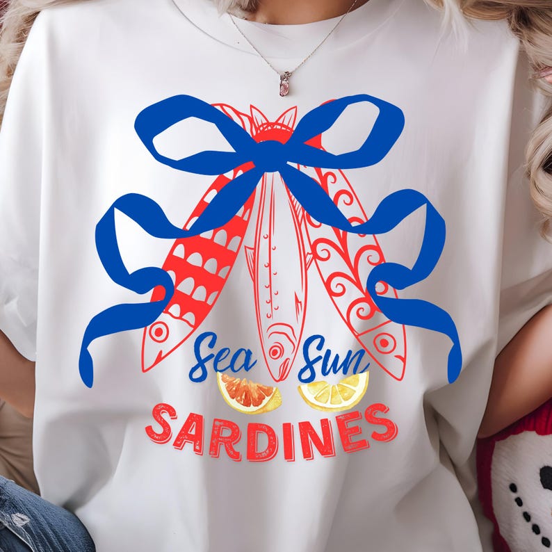 Puede incluir: Camiseta blanca con un lazo azul y una ilustraci&oacute;n de peces rojos y blancos. El texto "Sea Sun Sardines" est&aacute; impreso debajo de los peces. Dos c&iacute;tricos est&aacute;n debajo del texto.