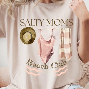 May include: A white t-shirt with the text "Salty Moms Beach Club" in brown lettering. The design includes a straw hat, a pink swimsuit, and a striped towel hanging on a clothesline.