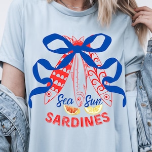 Puede incluir: Una camiseta azul claro con un gr&aacute;fico rojo y azul de tres peces atados con un lazo azul. El texto "Sea Sun Sardines" est&aacute; impreso debajo de los peces con dos rodajas de lim&oacute;n.