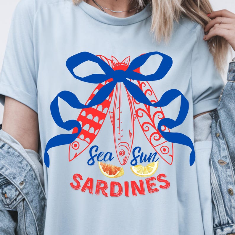 Puede incluir: Una camiseta azul claro con un gr&aacute;fico rojo y azul de tres peces atados con un lazo azul. El texto "Sea Sun Sardines" est&aacute; impreso debajo de los peces con dos rodajas de lim&oacute;n.