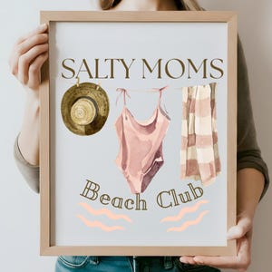 May include: A framed print with the text "Salty Moms Beach Club" in brown lettering. The print features a watercolour illustration of a swimsuit, a straw hat, and a striped towel hanging on a clothesline.