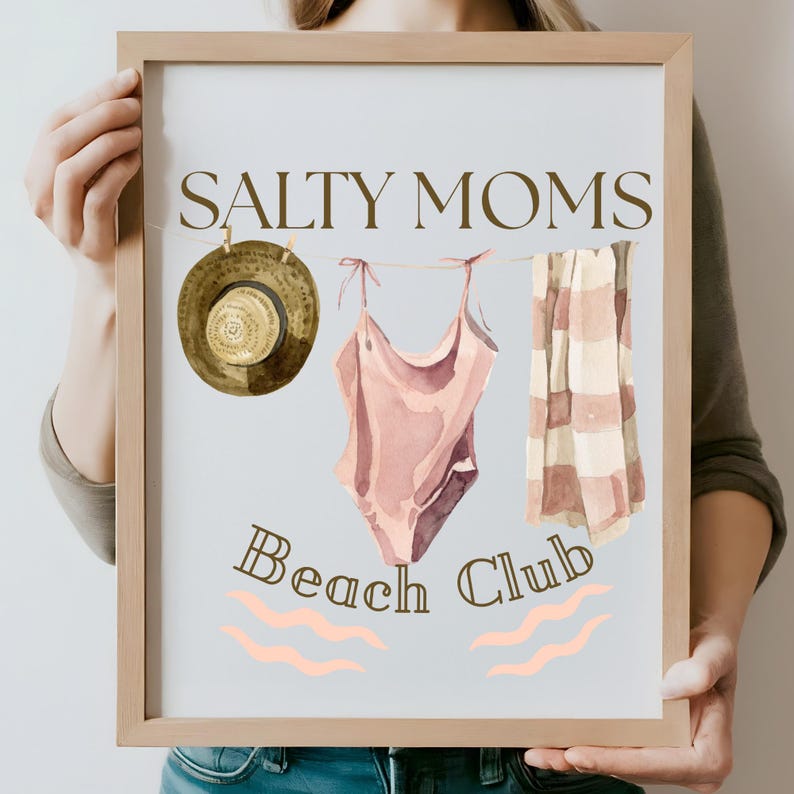 May include: A framed print with the text "Salty Moms Beach Club" in brown lettering. The print features a watercolour illustration of a swimsuit, a straw hat, and a striped towel hanging on a clothesline.