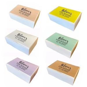 Puede incluir: Seis pequeñas cajas de regalo con tapas blancas y fondos coloridos. Cada caja tiene el texto "Olivia's First Birthday 11.10.2022" impreso en el fondo. Las cajas son de color rosa, amarillo, verde, morado, naranja y azul claro.