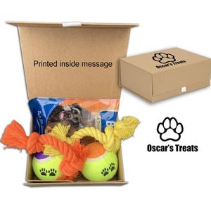 May include: A brown cardboard box with a printed message inside. The box is open and contains a dog toy set with two tennis balls, a rope toy, and a bag of dog treats. The box is labelled "Oscar's Treats" with a paw print logo.