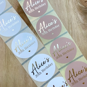 May include: Close-up of sticker rolls with a repeating design. Each round sticker features the name "Alice's" in a metallic gold script, along with "30th Birthday" and a small heart. The stickers are in shades of pink and light blue.