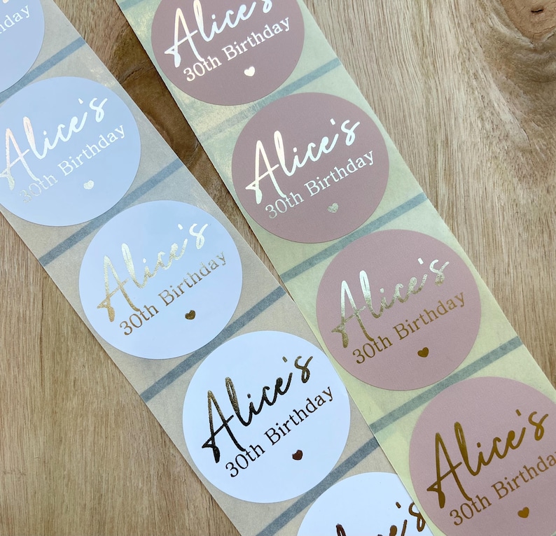 May include: Close-up of sticker rolls with a repeating design. Each round sticker features the name "Alice's" in a metallic gold script, along with "30th Birthday" and a small heart. The stickers are in shades of pink and light blue.