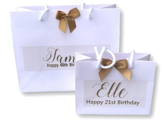 May include: Two white gift bags with gold bows and personalised labels. The larger bag says "Jam Happy 40th Bir" and the smaller bag says "Elle Happy 21st Birthday".