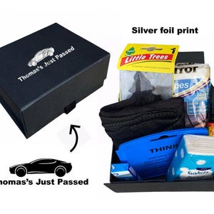 May include: A black gift box with a silver foil print of a car and the text "Thomas's Just Passed". The box is open and contains a variety of items, including a black cloth, a blue cleaning cloth, a pack of tissues, and a black marker.