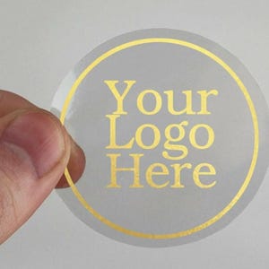 May include: A clear, circular sticker featuring a gold border and gold text reading "Your Logo Here." The sticker is held by fingers against a light grey background.