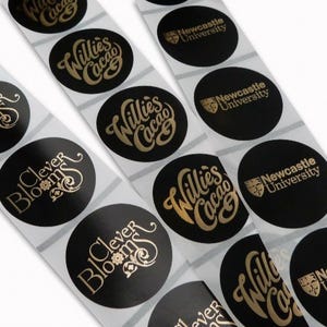 May include: A roll of black and gold round stickers with various designs. The designs include "Clever Blooms", "Willies Cacao", and "Newcastle University".