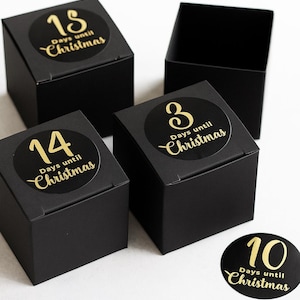 Puede incluir: Cajas cuadradas negras con pegatinas doradas que dicen "Días para Navidad". Las pegatinas muestran los números 18, 14, 3 y 10. Una caja está abierta, revelando su interior.