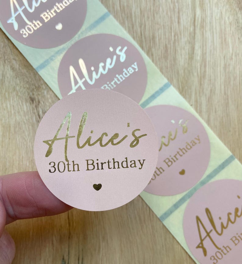 May include: A roll of pink stickers with gold foil text that reads "Alice's 30th Birthday". The stickers have a small heart symbol. The stickers are on a roll with a light blue and white striped backing.