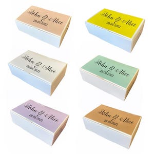 以下が含まれることがあります： 異なる色の蓋が付いた白いギフトボックスが6つあります。蓋の色は、ピーチ、イエロー、グリーン、パープル、ブラウン、ライトブルーです。各蓋には、「John & Alice」というテキストと「29.05.2022」という日付が黒で印刷されています。