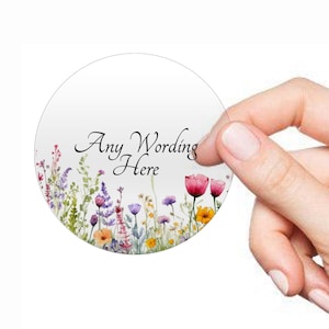 Peut inclure: Autocollant rond avec une illustration aquarelle d'un champ de fleurs sauvages dans des tons de rose, violet, jaune et vert. Le texte "Any Wording Here" est imprimé en noir au centre de l'autocollant.