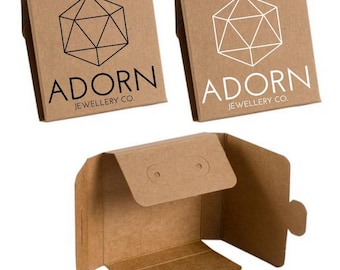 May include: Two brown cardboard boxes with a geometric logo and the text "Adorn Jewellery Co.". One box has a black logo and the other has a white logo. A third box is open and shows the inside of the box, which is also brown cardboard.